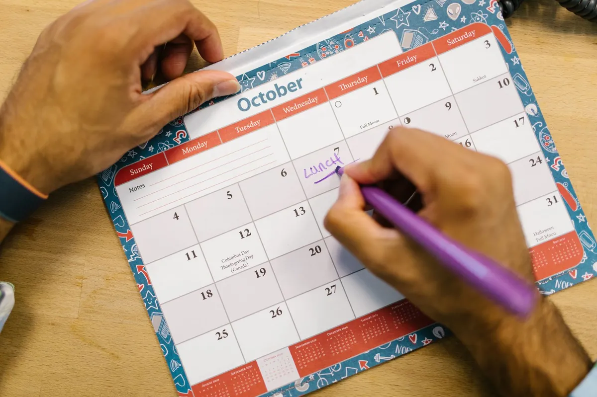 Close-up of a hand writing a lunch reminder on an October calendar with a purple pen.
