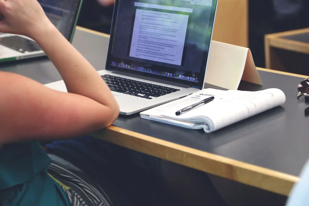 A student sits at a classroom desk, typing on a laptop while writing notes in a notebook.